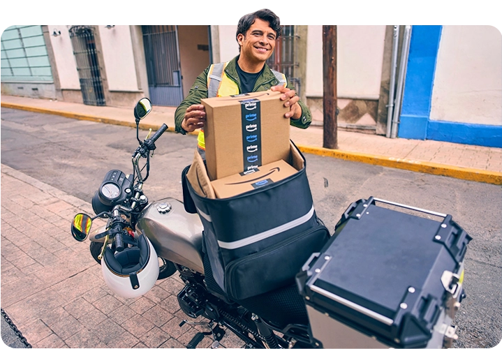 Repartidor con chaleco reflectante colocando paquetes de Amazon Prime en una bolsa de reparto térmica de color negro, sobre una motocicleta estacionada en una calle.